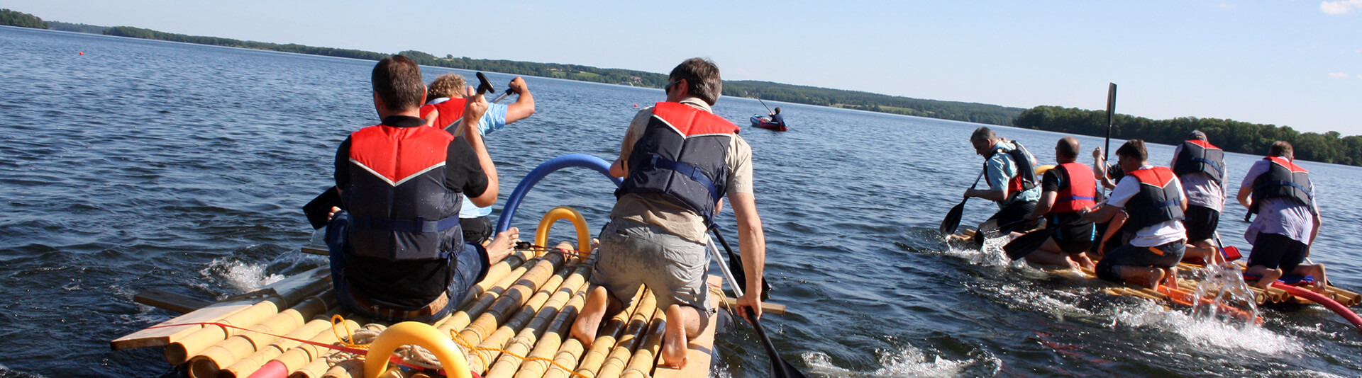 Mehrere Personen paddeln in zwei Floßen und einem Boot auf einem See